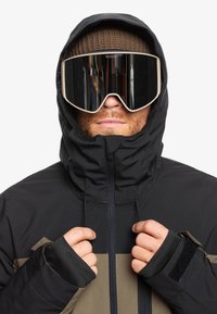 Doudoune noire et beige avec un col haut, fermeture éclair et trous pour les pouces. De grandes lunettes de ski miroir couvrent les yeux, portées avec un bonnet en tricot marron.
