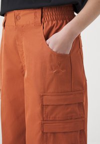 Orange cargo pants with an elastic waist, side pockets, and two large patch pockets on the thigh. Features embroidered logo detail.