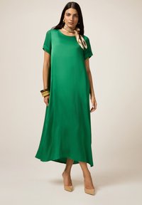 Abito maxi in satin verde con vestibilità rilassata, maniche corte e scollatura arrotondata, caratterizzato da un orlo asimmetrico e dettagli minimi.