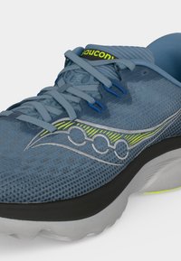 Gros plan d'une chaussure de running Saucony bleue avec des lacets bleu clair, un détail de logo jaune-vert et une semelle grise.