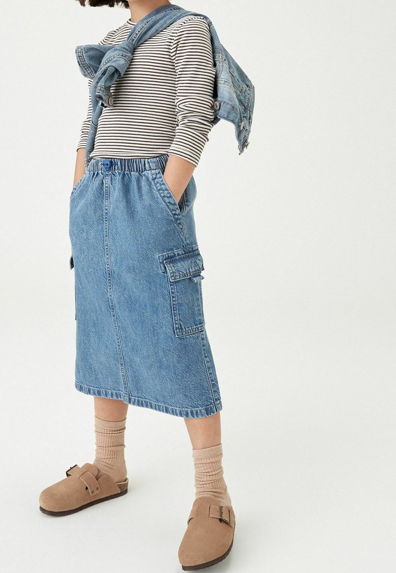 Enfant portant un t-shirt rayé à manches longues, une jupe en jean avec des poches, une veste en jean nouée autour des épaules, des chaussettes beiges et des chaussures marron sans lacets.