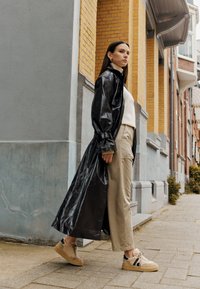 Black long trench coat with shiny finish, beige sweater, loose beige pants, and cream sneakers with fur texture and black accents.
