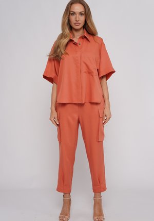 Frau trägt ein locker sitzendes kurzärmeliges rostfarbenes Hemd und passende Cargohosen, steht mit beigen Sandalen mit hohen Absätzen vor einfarbigem Hintergrund.