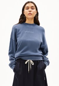 Sudadera de forro polar azul con cuello redondo acanalado, hombros caídos y una impresión de logo sutil. Combinada con pantalones negros con cintura ajustable.