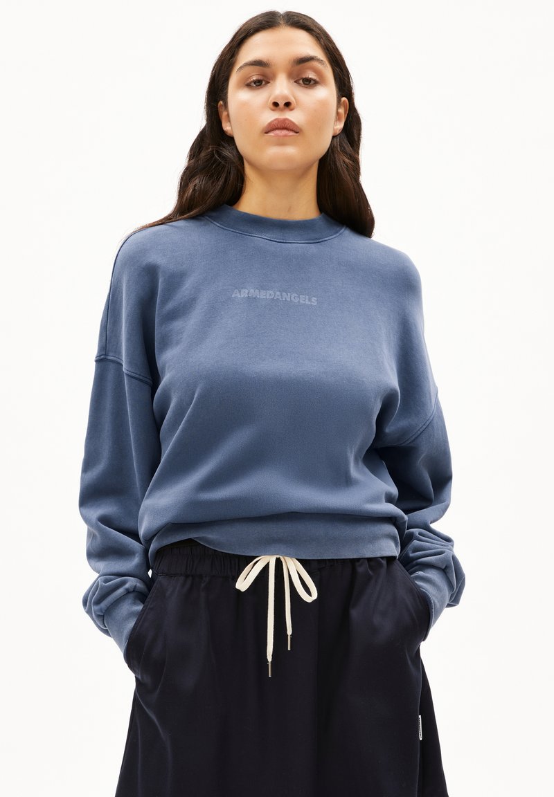 Sudadera de forro polar azul con cuello redondo acanalado, hombros caídos y una impresión de logo sutil. Combinada con pantalones negros con cintura ajustable.