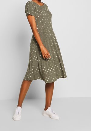 Vrouw draagt een olijfgroene jurk met korte mouwen met witte polkadots en witte sneakers, staand tegen een effen lichte achtergrond.