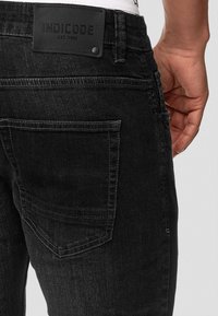 Svarta denimjeans med medelhög midja, med en läderpatch märkt 'INDICODE' på baksidan. Inkluderar två bakfickor.