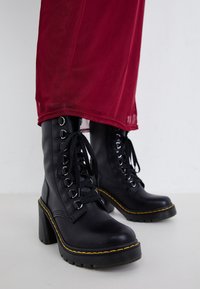 Black leather lace-up heeled boots with yellow stitching worn with a long red dress on a white background.