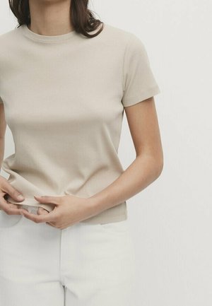 Femme portant un haut beige à manches courtes côtelé et un pantalon blanc, ajustant l'ourlet du haut avec les deux mains.