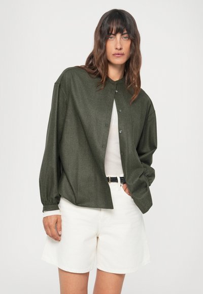 Cămașă verde închis, supradimensionată, cu nasturi, dintr-un material texturat, cu guler de tip bandă și mâneci lungi, purtată cu pantaloni scurți albi pentru un contrast.