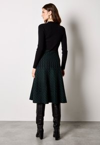 Donna che indossa un top nero a maniche lunghe a coste, gonna a linea A verde con motivo, stivali neri al ginocchio e grandi orecchini a cerchio, in piedi contro una parete bianca.