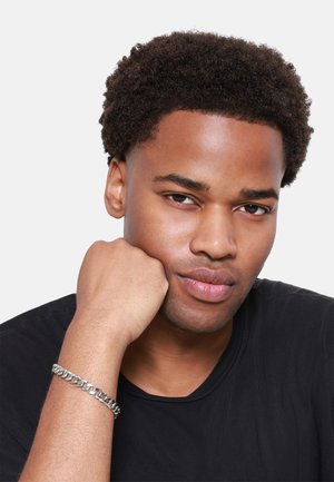 Bracelet en chaîne argentée porté au poignet, avec des motifs entrelacés. Le porteur a les cheveux courts et bouclés de couleur sombre et porte un t-shirt noir.