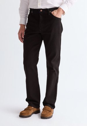 Homme portant un jean noir droit, une chemise blanche et des mocassins en daim marron, une main dans la poche devant un fond uni.