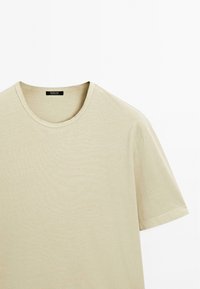 T-shirt in cotone beige con scollatura rotonda e maniche corte. Presenta una texture liscia e un design minimale senza motivi o dettagli visibili.