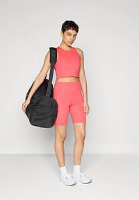 Ροζ κοντό τοπ και ταιριαστά ποδηλατικά σορτς, σε συνδυασμό με μαύρη τσάντα duffel. Το μοντέλο φοράει λευκά αθλητικά παπούτσια και κάλτσες αστραγάλου. Καθαρό φόντο.