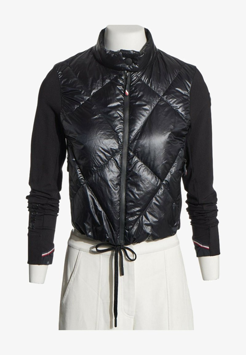 Doudoune sans manches noire matelassée avec col montant et fermeture éclair devant, portée sur des manches longues noires, associée à un pantalon blanc taille haute.