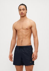 Shorts de bain bleu marine en matière légère avec une taille élastique et un cordon de serrage. Présente un logo sur le devant et une texture sur les côtés.