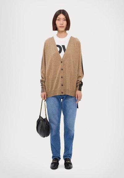 Cardigan marrone con scollo a V, con bottoni frontali e maniche lunghe, indossato sopra una t-shirt bianca con stampa nera, abbinato a jeans blu e una borsa nera.