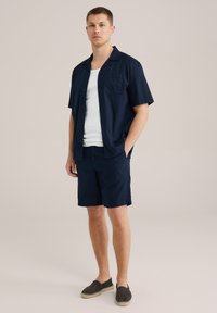 Camisa de lino navy de manga corta combinada con pantalones cortos a juego y alpargatas negras. Cuenta con un bolsillo en el pecho y un diseño de corte relajado.