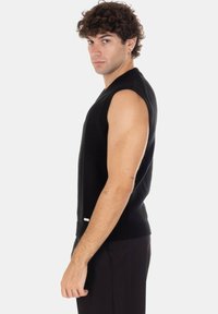 Giovane uomo con capelli castani ricci, indossa un maglione nero senza maniche e pantaloni neri, in piedi di profilo su uno sfondo bianco.