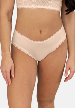 Frau trägt hellbeige Unterwäsche mit Spitzenbesatz und Spitzen-Bralette, mit Bauchnabelpiercing und Tätowierungen am linken Arm, stehend vor weißem Hintergrund.