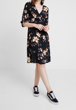 Kvinde iført en sort knælængde kjole med orange og hvide blomsterprint, kombineret med sort-hvide sneakers, stående mod en ensfarvet baggrund.