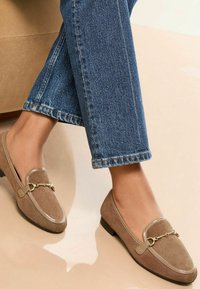 Beige mokkasiner i semsket skinn med en liten hæl, med metallisk aksent og dekorative sømmer, kombinert med mørkeblå denim.