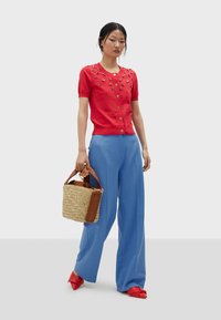 Cardigan court rouge à manches courtes avec des embellissements décoratifs, associé à un pantalon bleu ample et un sac en paille tressé avec des accents marron. Talons rouges.