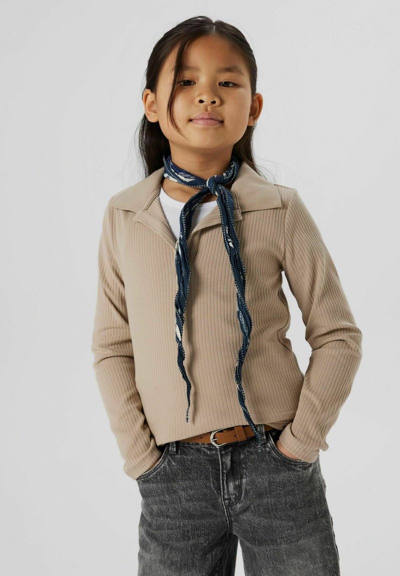 Beige ribbad långärmad topp med krage och en marinblå mönstrad scarf knuten runt nacken, i kombination med grå denimbyxor och ett brunt bälte.