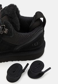 Baskets basses noires avec une tige en daim et en maille, une semelle en caoutchouc texturée, et deux lacets noirs à côté.