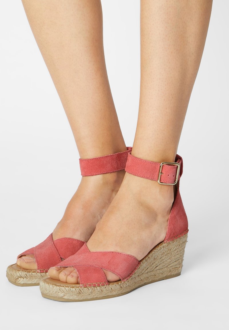 Piedi che indossano sandali con zeppa in suede color corallo e cinturini alla caviglia, con suole di piattaforma in juta intrecciata su uno sfondo bianco.