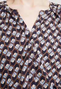 Blusa marrone con un motivo esagonale in beige e blu chiaro. Ha una scollatura a V e un tessuto morbido e testurizzato.