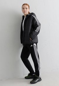 Svarte pufferjakker med horisontale sømmer, hvite striper på ermene, sammen med svarte treningbukser med hvite detaljer og en hvit skjorte.