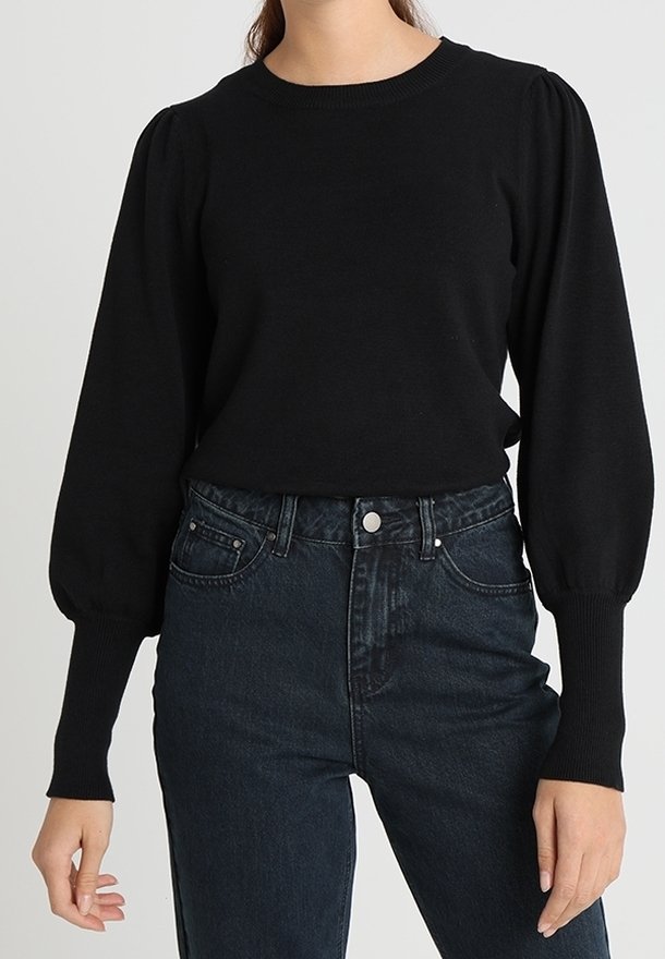 Femme portant un pull noir à manches longues avec des épaules bouffantes, rentré dans un jean bleu foncé taille haute.