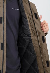 Alyvos žalumo spalvos išorinis švarkas su juodu pagalvėlėmis, užsegimas užtrauktuku ir Velcro akcentais; dėvimas ant tamsiai mėlynos marškinėlių su logotipu.