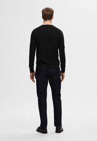 Pull noir à manches longues avec poignets côtelés ; porté avec un jean bleu foncé et des chaussures noires ; design simple, silhouette ajustée, détails minimalistes.