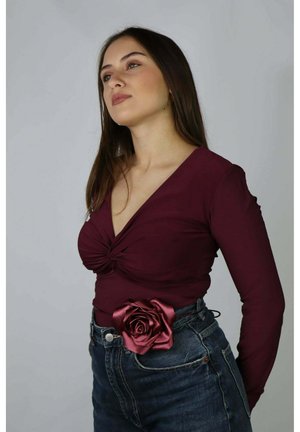Młoda kobieta ubrana w bordową bluzkę z długim rękawem z wiązanym przodem, wysokim stanem dżinsy oraz satynowy bordowy pasek z różą.