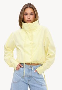 Femme portant une veste jaune à col montant, boutonnée, avec des cordons de serrage et un jean bleu clair, face à l’avant, les mains dans les poches.