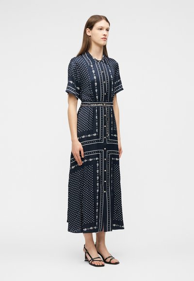 Γυναίκα που στέκεται, φοράει ένα midi φόρεμα σε ναυτικό μπλε χρώμα με κουμπιά, εμπριμέ, με κοντά μανίκια και μαύρες δερμάτινες πλατφόρμες με λουριά, σε απλό φόντο.