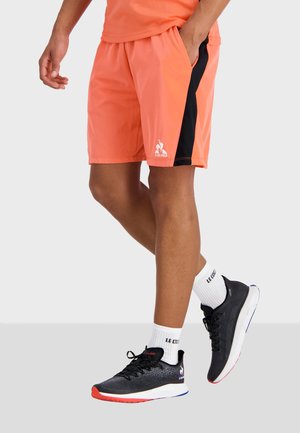 Shorts de sport orange clair avec des panneaux latéraux noirs, taille élastique et poches. Accompagnés de chaussures de sport noires et de chaussettes blanches.