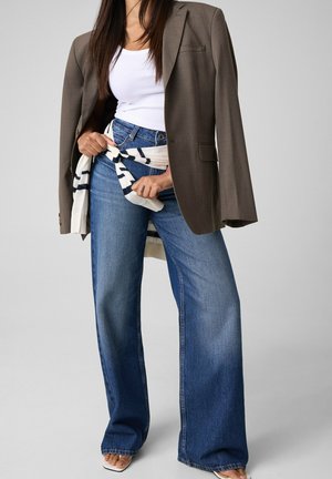 Donna che indossa un blazer marrone sopra una canotta bianca con un maglione a righe legato intorno alla vita e jeans blu a gamba larga.