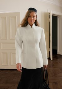 Balta pritaikyta striukė su aukštu apykakle, struktūruotais pečiais ir šoninėmis kišenėmis, derinama su ilgu juodu plisuotu sijonu ir juoda rankine.