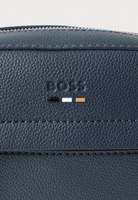 Tengerészkék, texturált bőr táska dombornyomott "BOSS" logóval és három kis díszcsíkkal fekete, fehér és barna színekben. Cipzáros felső záródás.