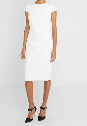 Vrouw die een nauwsluitende, knie-lange witte jurk draagt met cap mouwen en zwarte hoge hakken met strikjes bij de enkels.