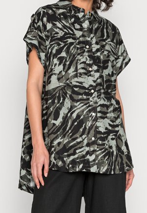 Femme portant une chemise à manches courtes boutonnée avec un imprimé zèbre abstrait en noir, gris et vert olive, associée à un pantalon noir.