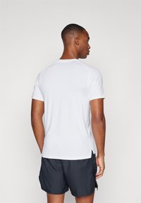 White short-sleeve athletic shirt with a crew neck, featuring textured fabric and side slits, worn with dark shorts.