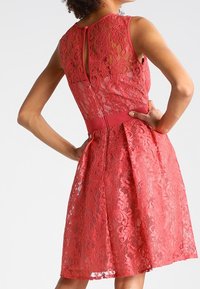 Korall spetsklänning med figurnära liv, utsvängd kjol och bakdragkedja. Har intrikata blommönster och ett solid midjeband.
