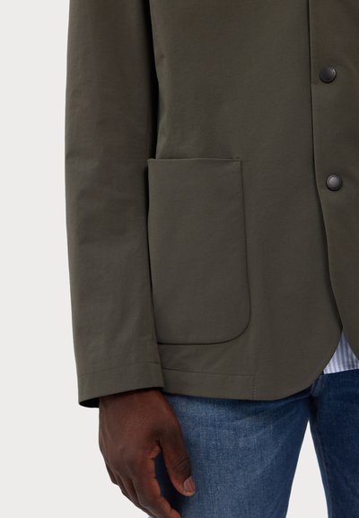 Olive green jacket with a patch pocket, black buttons, and a soft texture. Worn over a blue striped shirt and denim jeans.