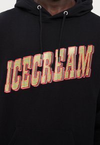 Schwarzer Hoodie mit der Aufschrift "EISCREME" in großen, gelben, strukturierten Buchstaben, die rot umrandet sind. Mit einer Fronttasche und verstellbaren Kordeln.