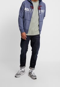 Navy zip-up hoodie med vit och röd text "Hollister California", grå t-shirt, mörka jeans och grå sneakers med vita snörningar.
